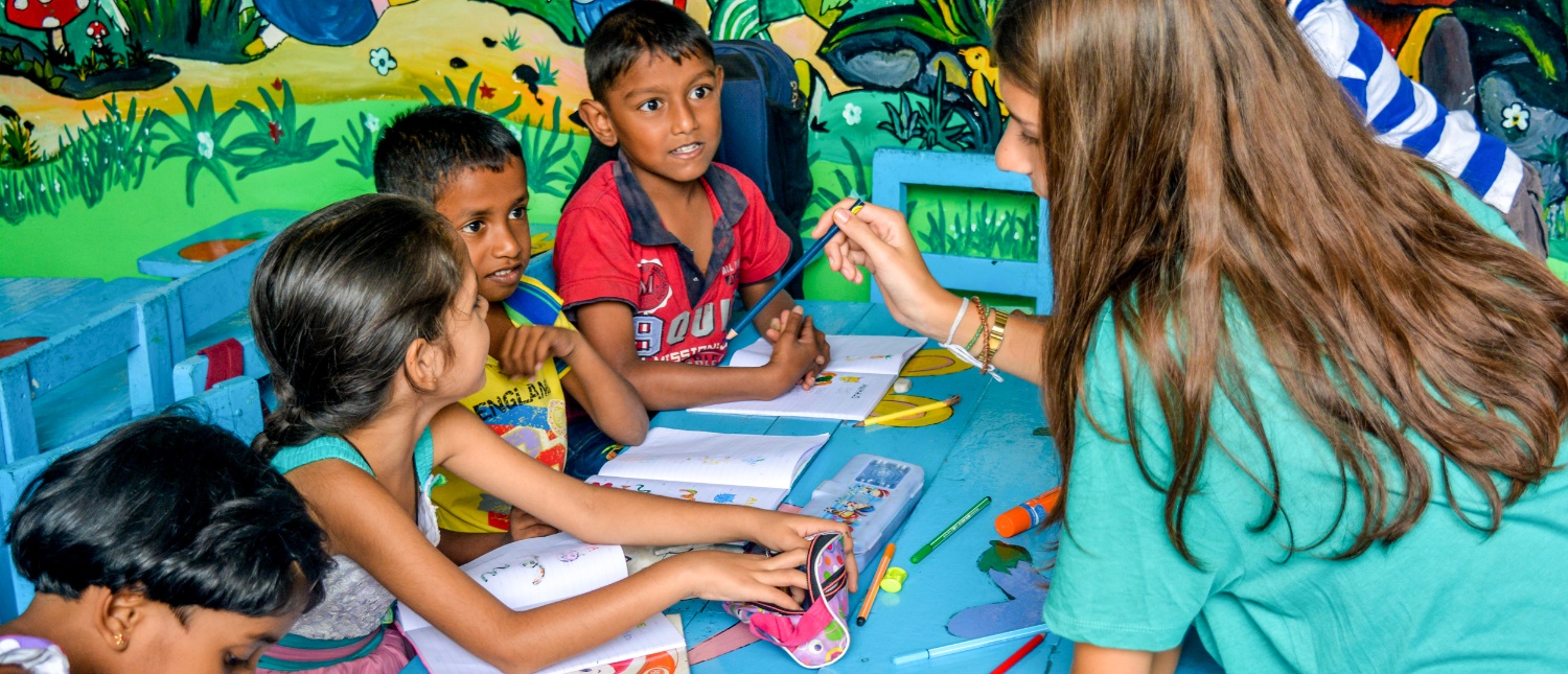 A volunteer teacher guides young children drawing at a blue table in a colorful Sri Lankan preschool.